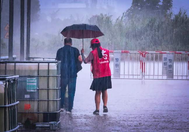 暴雨中,一位身穿红色小马甲,梳着马尾辫的志愿者姑娘,撑伞扶送一位